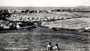 Old black and white image of touring caravans in the field behind the village with the sea in the back ground.