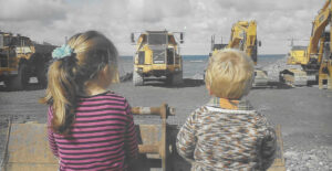 Two children sit watching diggers and dumber trucks working on the beach