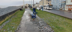 Beach stones scattered across the prom by a bench after a storm