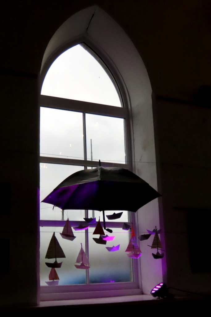 Paper boats hang from a broken umbrella in a chapel window