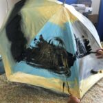 A person paints black paint on to any umbrella to create a stormy sky.
