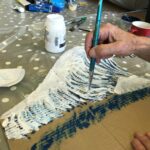 A person's hand holds a paint brush as they paint detail on to a cardboard wave.