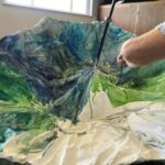 A person's arm holds a paint brush as they paint stormy water on to an old umbrella.