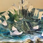 White paper boats and white miniature laundry arranged in an up turned umbrella that is painted like a stormy sea