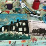 A painting of houses next to a stormy sea takes shape on a table with a cup of tea