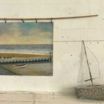 A painting of a boat on the beach hangs on a white wall beside a newspaper model boat