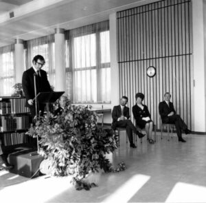 A black-and-white photo of an exhibition opening in an open space in a library building, which is an open space with low, shoulder-height shelves, relatively low ceilings, and floor-to-ceiling windows. A man is giving a lecture on a podium that has a large plant in front of it. To his left, 3 people are watching with their heads turned up towards him: 2 men, and in between them is a woman with a beehive hairstyle.