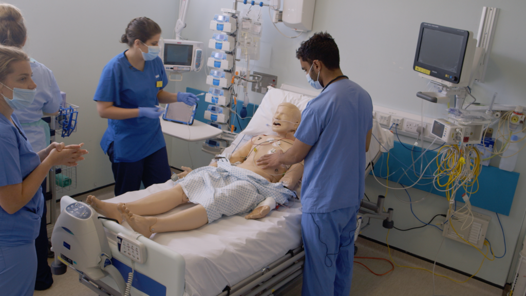 Four health professionals wearing blue scrubs stood around a training mannequin, taking part in a simulation-based training exercise at a hospital ward.