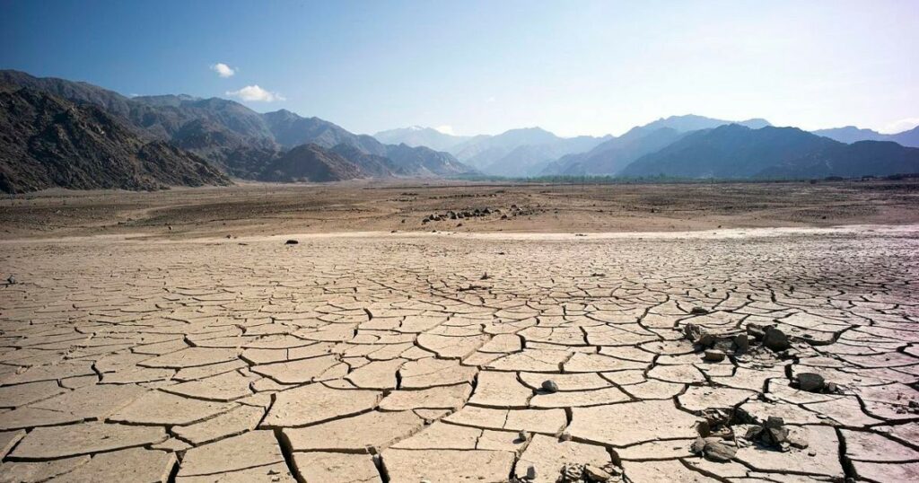 A wide, arid landscape of cracked, sun‑baked earth stretching toward distant rocky mountains under a clear blue sky, illustrating severe drought and land degradation.