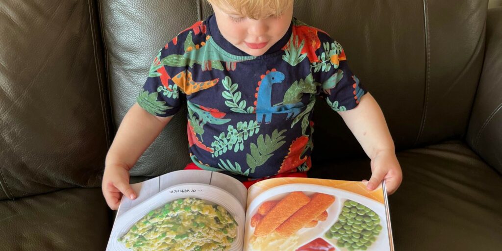 Young child engaging with a picture book featuring images of meals with vegetables, supporting early food familiarity.
