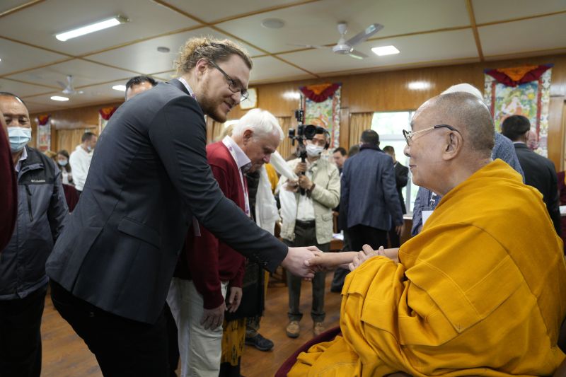 A man in a suit with glasses, a beard and pony tail shakes the hand of a man dressed in a yellow robe