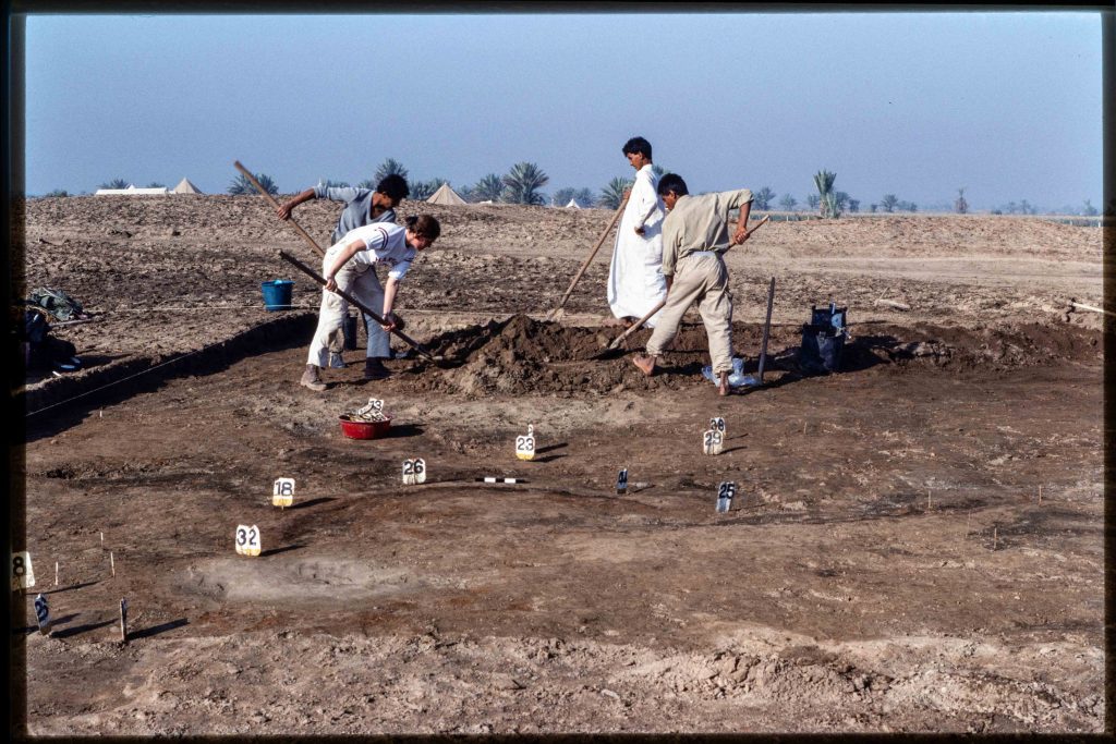 Several men with shovels work in the background at an archaeological site; in the foreground there are several signs on the ground with numbers pointing to feature discovered through excavation
