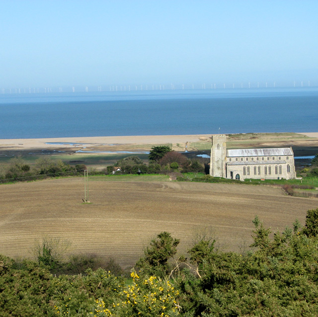 Photograph of St Nicholas church with a ploughed field and trees in the foreground in front of the church, and saltmarshes and sea in the background behind the church.
