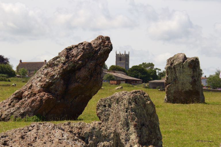 Photo of Stanton Drew Church square tower as seen behind a three standing stones in a field