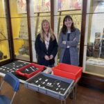 Lynn Museum staff Jan Summerfield and Dayna Woolbright standing in front of the medieval object cases at Lynn Museum and behind a table with trays of pilgrim badges
