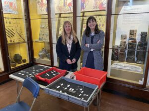 Lynn Museum staff Jan Summerfield and Dayna Woolbright standing in front of the medieval object cases at Lynn Museum and behind a table with trays of pilgrim badges