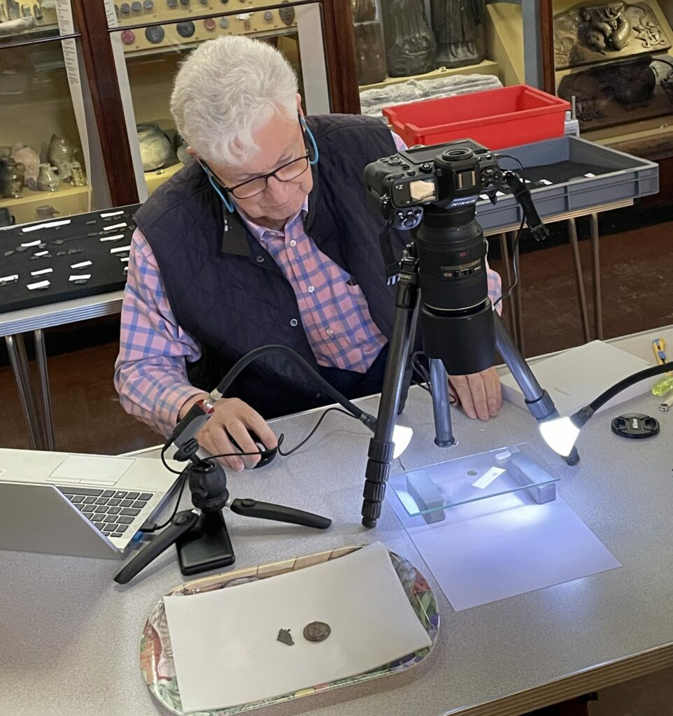 Rod Trevaskus sitting in front of medieval object cases at Lynn Museum. Rod is sitting at table photographing pilgrim badges. On the table to one side of Rod is a laptop. In front of Rod there is a tabletop tripod holding a camera which is pointing downwards towards a pilgrim badge mounted on two blocks supporting a clear sheet, all placed over a sheet of white paper. Two pilgrim badges are placed in a tray on white paper on the table also.