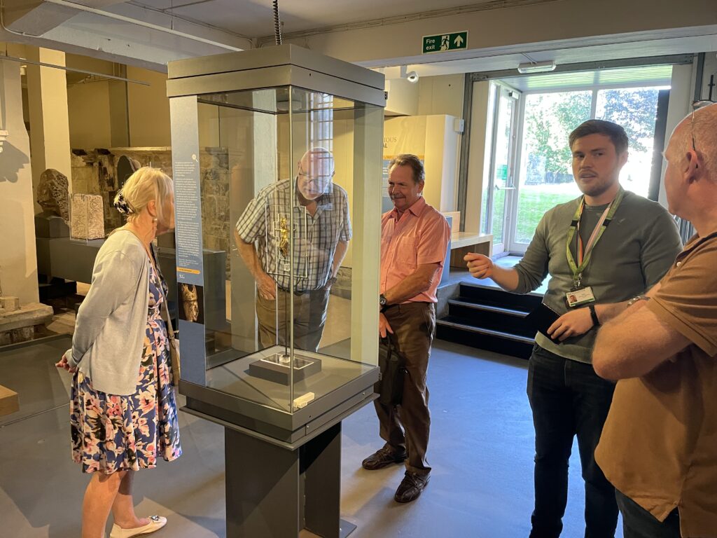 3 citizen scientists are standing around a display case featuring a gold medieval figurine of Christ on a crucifix. To the right side of the group, Harry Ellerd-Cheers, Associate Collections Curator is talking to another citizen scientist.