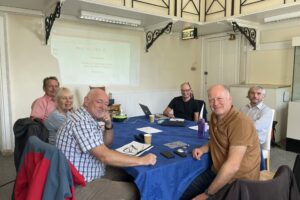 A group of 6 people sitting around a table covered with a blue cloth, archaeological objects, papers and a laptop. The group are looking towards the camera and smiling.