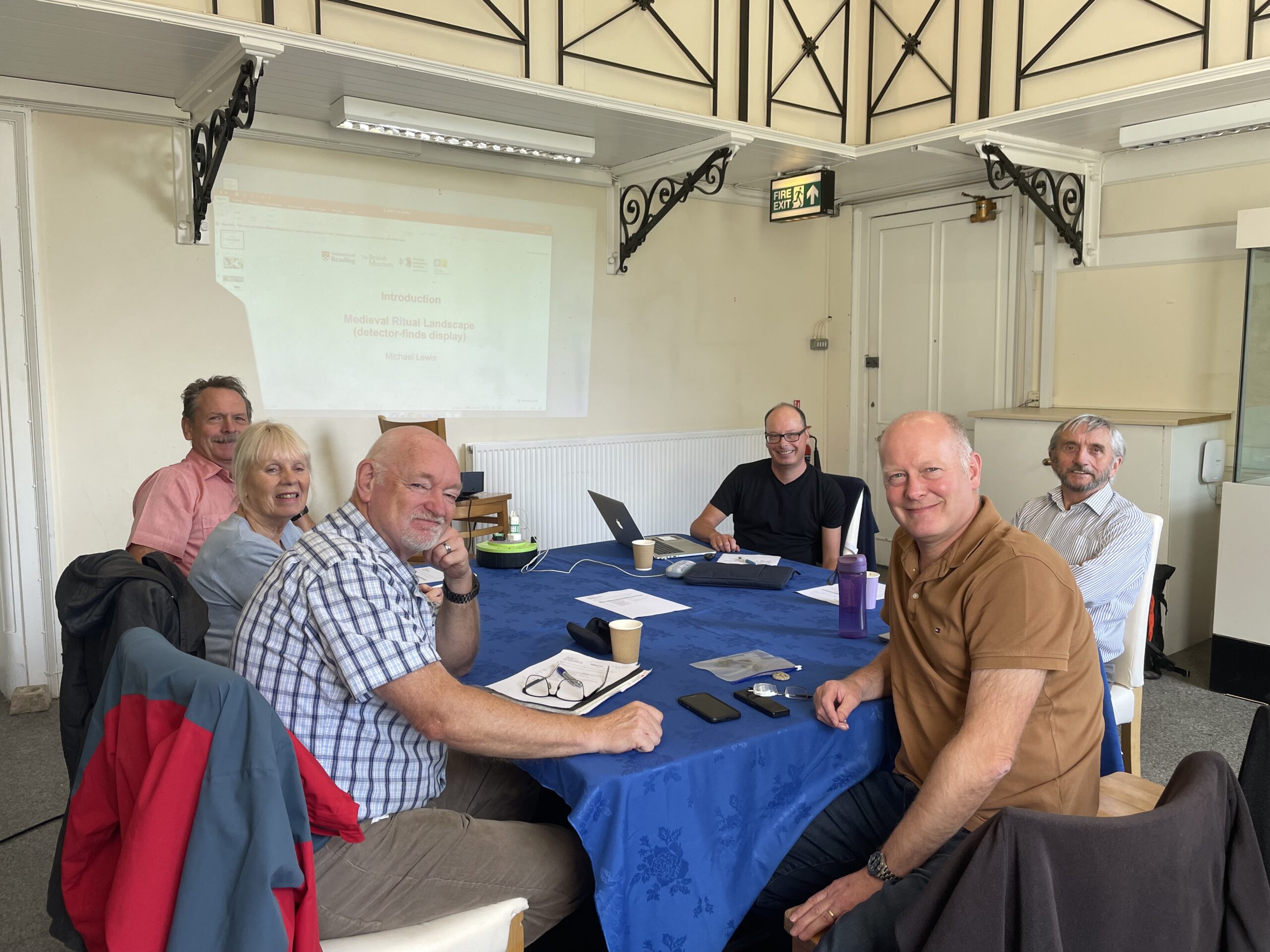 A group of 6 people sitting around a table covered with a blue cloth, archaeological objects, papers and a laptop. The group are looking towards the camera and smiling.