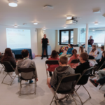 Professional male archaeologist talks through presentation to a group of seated children - members of Norwich Young Archaeologists' Club