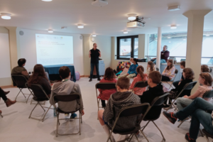 Professional male archaeologist talks through presentation to a group of seated children - members of Norwich Young Archaeologists' Club