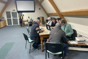 Six men sit around a table in the foreground, discussing lead objects, while two discuss metal analysis in the background