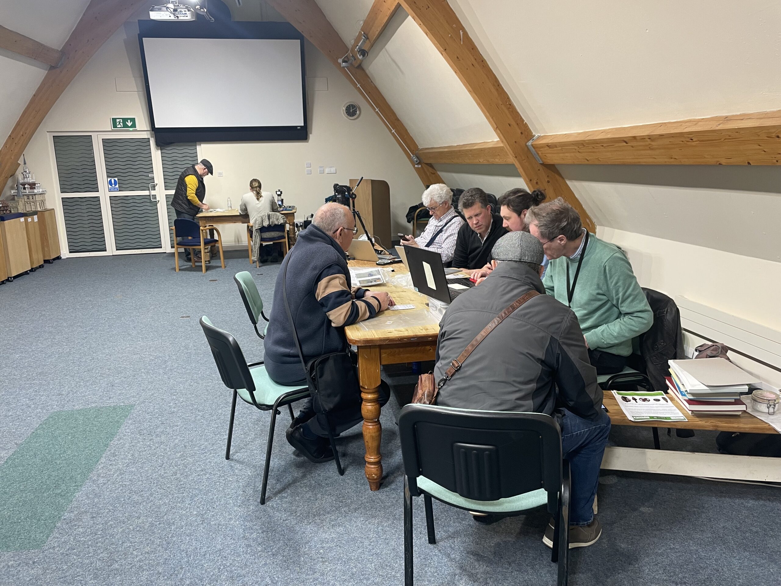 Six men sit around a table in the foreground, discussing lead objects, while two discuss metal analysis in the background