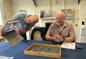One male standing holding a laptop over a table. There is another male seated at the table. A third male can be seen on the laptop screen. 2 are looking at a wooden tray containing 5 small artefacts on the table while the man holding the laptop is looking at the laptop screen