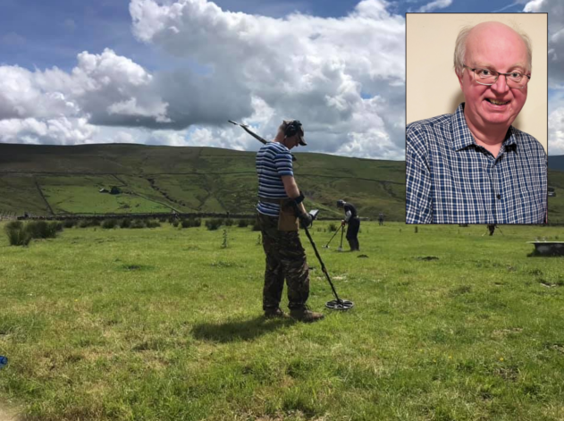 A man wearing a baseball cap, over ear headphones, a striped t shirt and camouflage pattern trousers holding a metal detector in a field with a hill in the background. Another person is detecting in the background. In the top right corner is an inset head shot of a smiling white man with white hair wearing glasses and a blue check pattern shirt