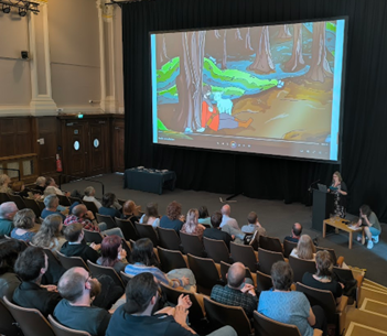 A group of people seated in rows in a lecture theatre with a large screen showing an animation screenshot of a white man with a brown beard and brown hair wearing a red long sleeved top, light orange belt, grey leggings and orange boots. The man sits on the floor leaning against a tree in a forest. A lamb stands next to the man.
