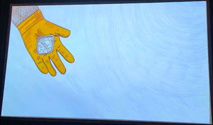 A light blue screen. In the top left hand corner is a cartoon hand wearing a yellow-orange glove and part of a grey arm decorated with a criss-cross pattern. The hand is holding a cartoon style lozenge shaped object showing the ‘Agnus Dei’ (Lamb of God) motif