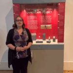 A white female with blonde hair, wearing glasses, a black cardigan, purple patterned top and black trousers standing in front of a display case with a red panel and text saying "THE MEDIEVAL RITUAL LANDSCAPE PROJECT". There are various small objects on grey mounts in the display case.