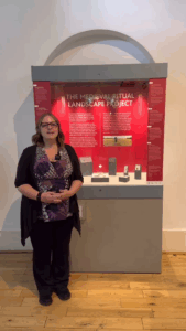 A white female with blonde hair, wearing glasses, a black cardigan, purple patterned top and black trousers standing in front of a display case with a red panel and text saying "THE MEDIEVAL RITUAL LANDSCAPE PROJECT". There are various small objects on grey mounts in the display case.