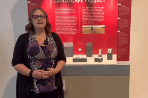 A white female with blonde hair, wearing glasses, a black cardigan, purple patterned top and black trousers standing in front of a display case with a red panel and text saying "THE MEDIEVAL RITUAL LANDSCAPE PROJECT". There are various small objects on grey mounts in the display case.