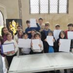 A group of children stand behind a table inside a church foyer room. There is a roll of paper with a large church plan diagram on the table and the children are holding up smaller paper display plans