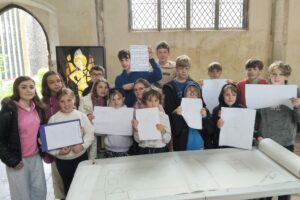 A group of children stand behind a table inside a church foyer room. There is a roll of paper with a large church plan diagram on the table and the children are holding up smaller paper display plans