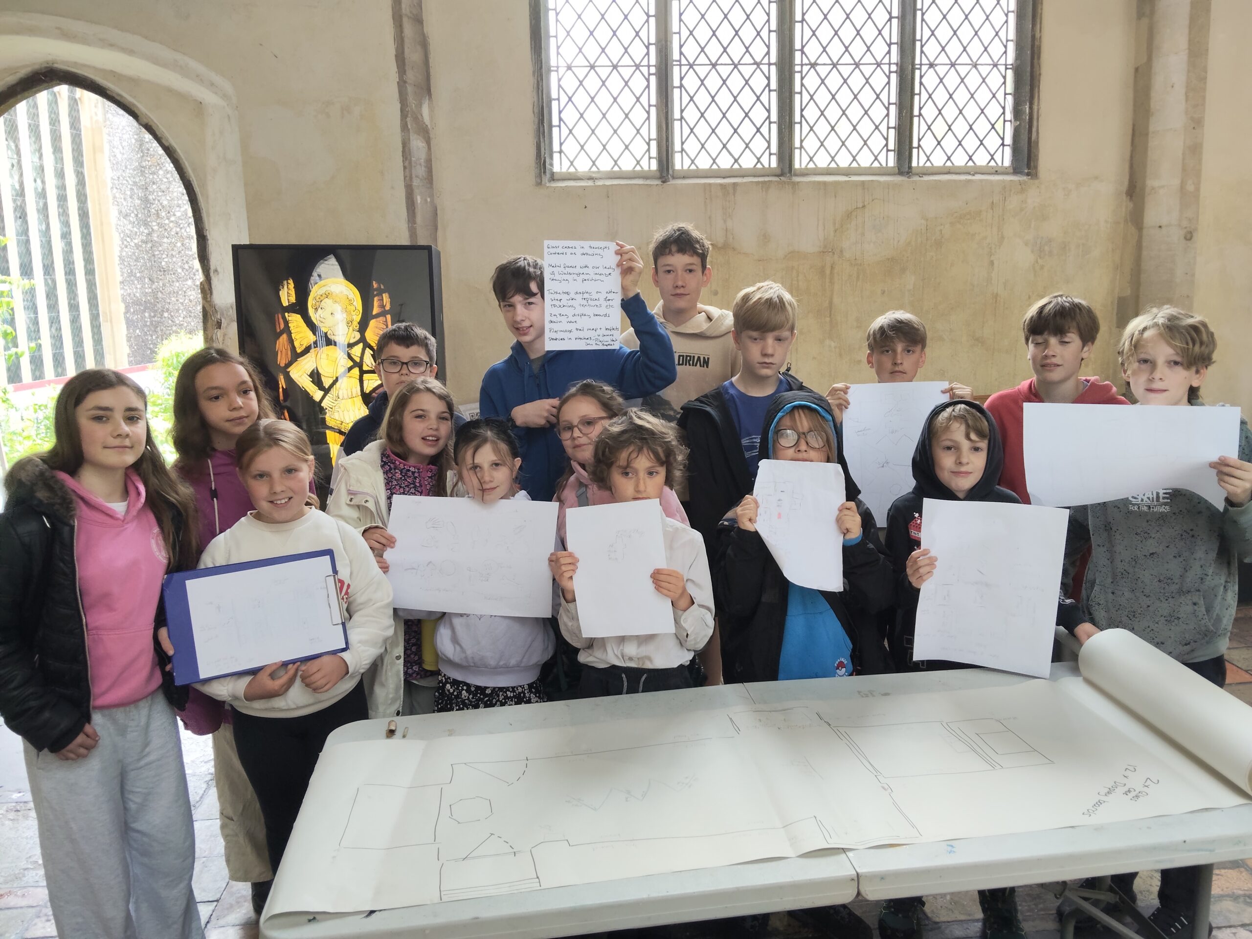 A group of children stand behind a table inside a church foyer room. There is a roll of paper with a large church plan diagram on the table and the children are holding up smaller paper display plans