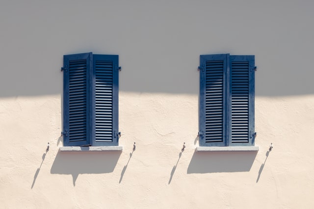 Two blue shutters cover windows on a white wall basked in sun. Extreme weather.