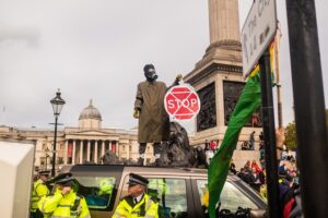 A climate activist wearing a gas mask and holding a stop sign stands on top of a parked car in Trafalgar Square. Police Officers surround the vehicle.
