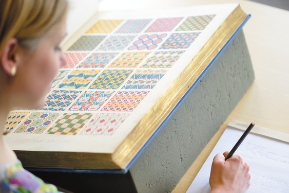 A woman makes notes as she looks at a large book of colourful patterns in an archive