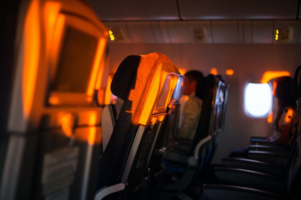 A row of seats on an airplane. Sunlight shines on the back of the seats. Two passengers are seated at the end of the row next to the windows.