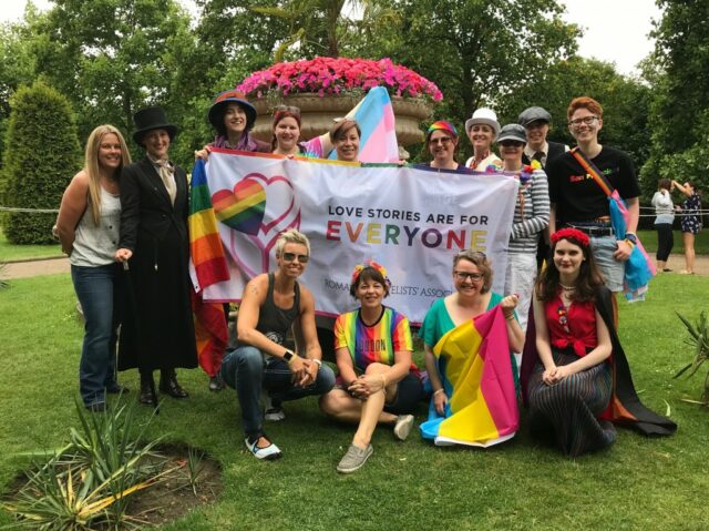 Members of the Romantic Novelists' Association pose with a banner depicting a rainbow heart and reading 'love stories are for everyone'. They are all smiling!