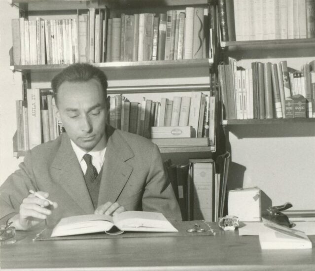 A black and white photograph of the writer Primo Levi sat at a desk reading a book in 1960. He is wearing a suit and tie. Behind him are shelves filled with books.