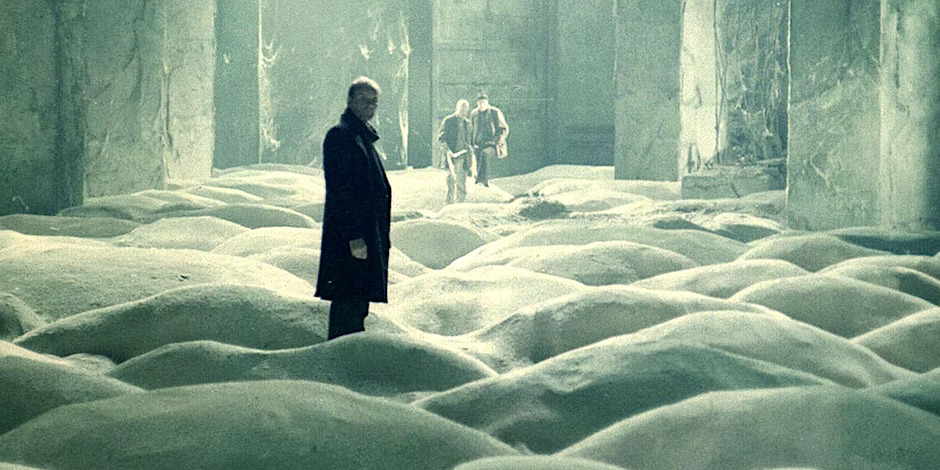 A still from Andrei Tarkovsky's film Stalker, showing a man standing in the foreground, facing the camera, in a snowy scrubland, with two men in the background looking at him.