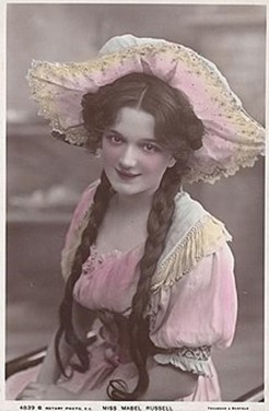 A picture of Mabel Philipson as a Gaiety Girl. Her hair is in long plaits. She is wearing a dress with lace trim and a broad brimmed sun hat.