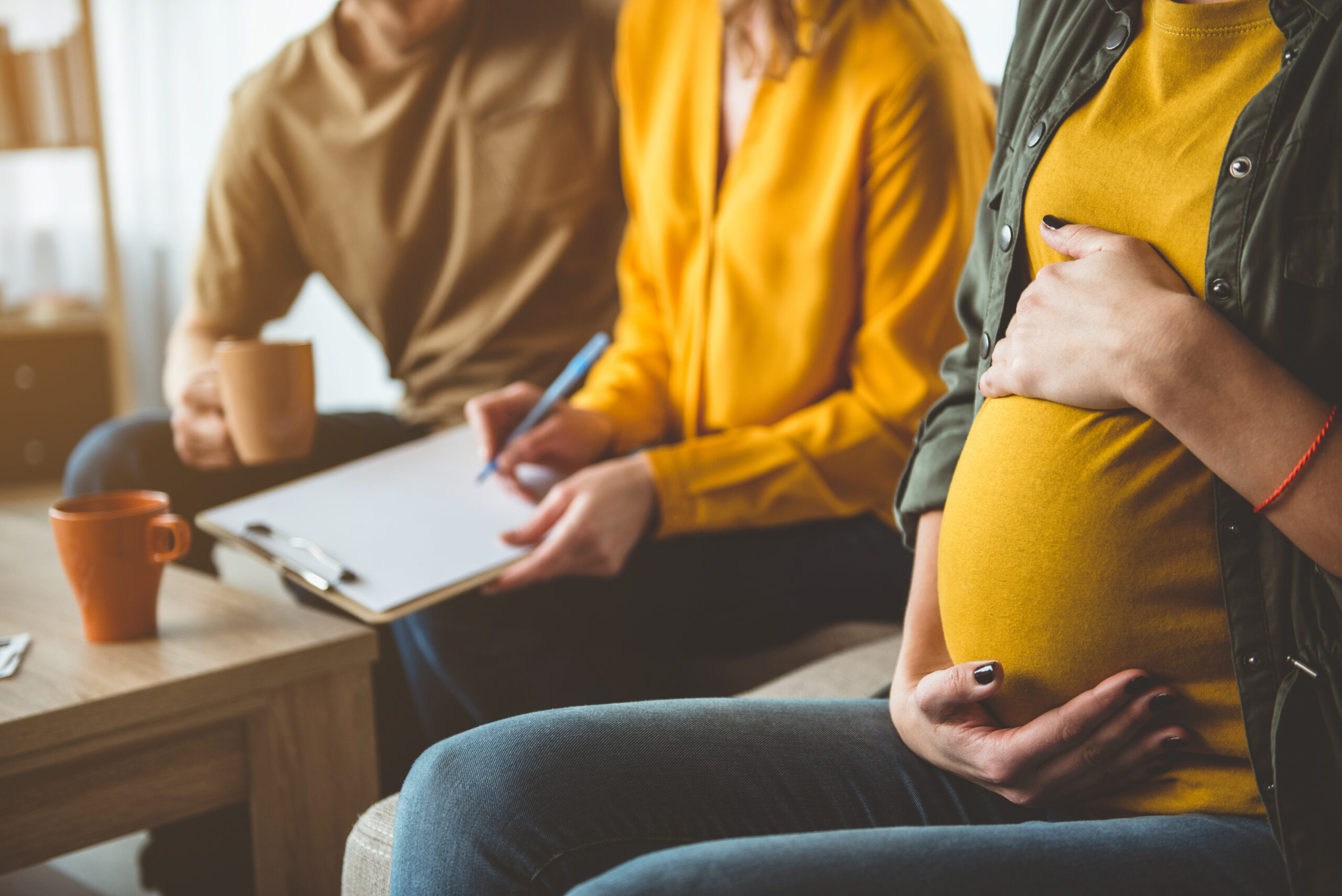 Focus on pregnant woman with hands on her abdomen. Married couple is putting signature on document while sitting on background. Surrogate motherhood concept
