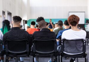 Teenagers in a large classroom. The view is of three students in the back row, paying attention to the teacher at the front.