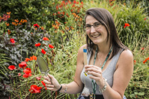 Soil scientist Sarah Duddigan holds a trowel, tea bag and marker stick. She is surrounded by vegetation and flowers and is smiling.