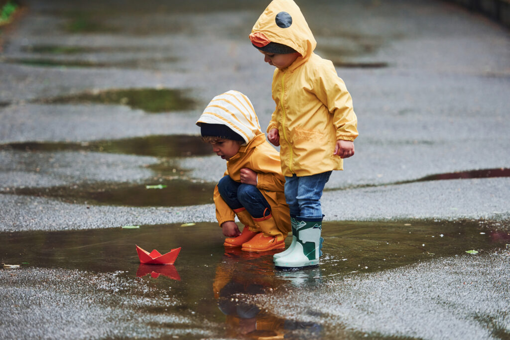Two kids in yellow waterproof cloaks and boots playing with handmade paper boat outdoors after the rain together.